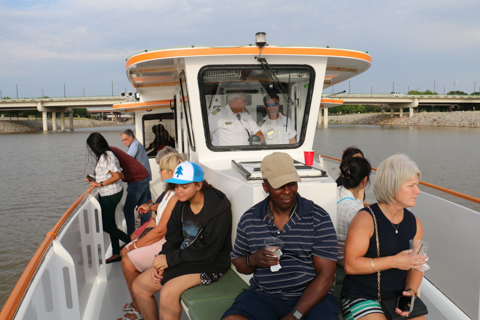 Group of individuals sitting on dock of Oklahoma River Cruiser while two captains are seen through the window steering.