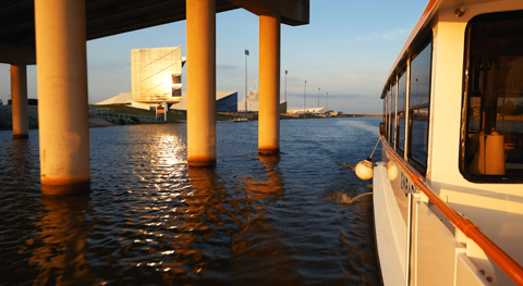 Oklahoma River Cruiser driving under the I-40 bridge with Oklahoma City Boathouse District in the background on a sunny evening.