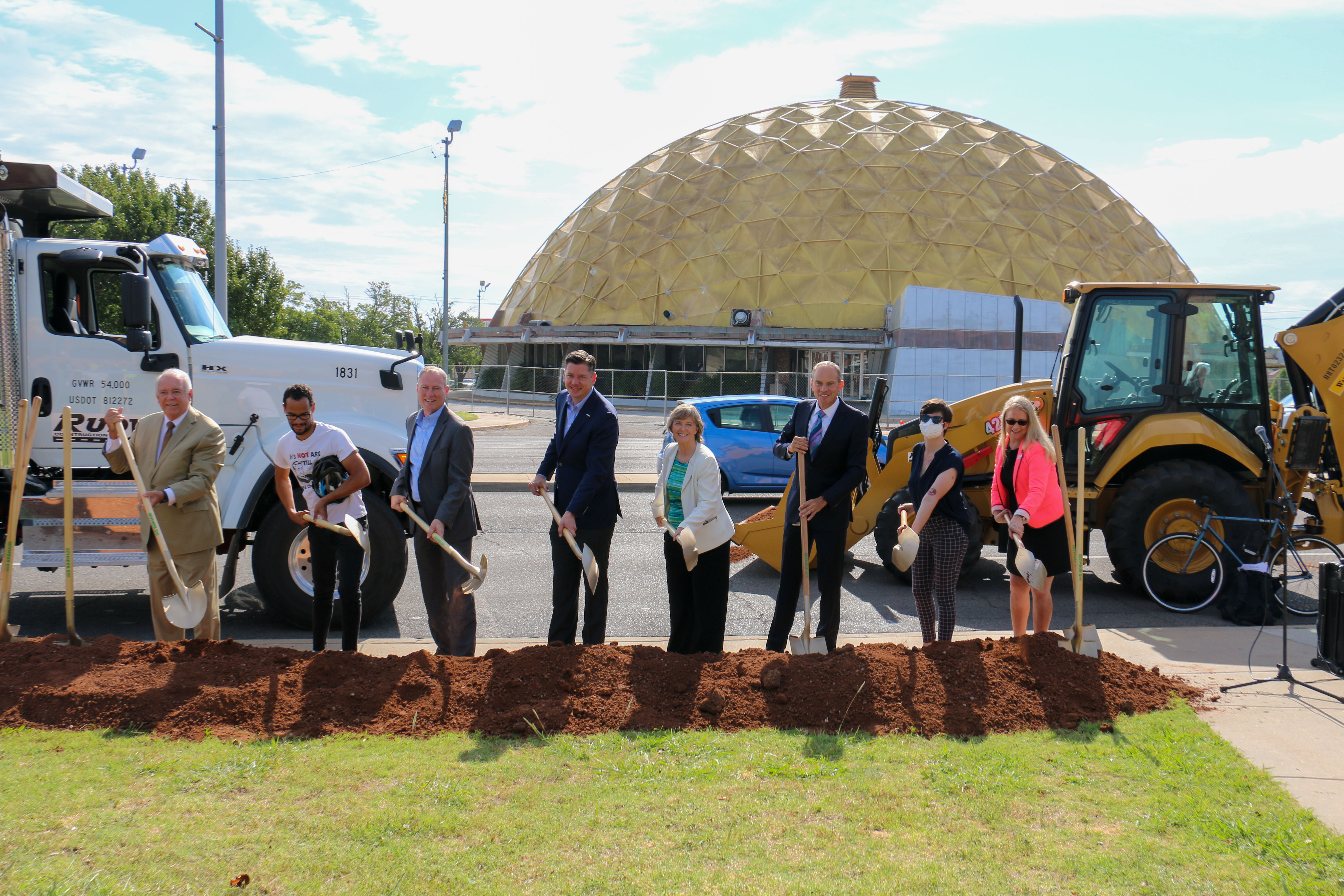 City Officials holding shovels in front of a pile of dirt on 23rd & Classen with Gold Dome and construction equipment in the background.